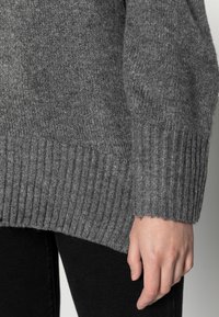 Nahaufnahme einer Hand, die seitlich neben einer Person ruht, die einen grauen Strickpullover und schwarze Hosen trägt.