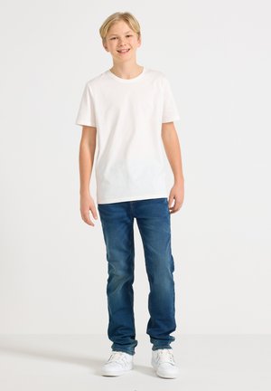 Lyshåret dreng står og smiler, iført en ensfarvet hvid T-shirt, blå jeans og hvide sneakers, foran en ensfarvet lys baggrund.