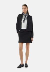 Zwarte cropped jas met twee voorzakken, witte geribde top en zwarte rok; geaccessoireerd met een patroon-sjaal en zwarte loafers.