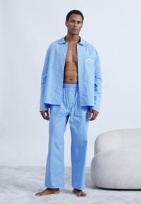 Σετ πιτζάμες με μπλε ρίγες, που περιλαμβάνει πουκάμισο με κουμπιά και λογότυπο στο στήθος, καθώς και παντελόνι με ελαστική μέση, κατασκευασμένα από ελαφρύ ύφασμα.