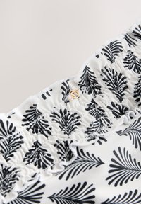 Tessuto bianco con motivi fogliari neri, caratterizzato da un dettaglio di balza arricciata ed elastica e da una piccola fibbia decorativa dorata.