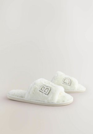 Biele plyšové papuče s chlpatou textúrou, so zažiarivými kamienkami na remienkoch a mäkkou, polstrovanou podrážkou.