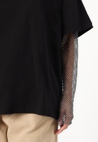 Melna T-krekls ar īsām piedurknēm, kura piedurknes izgatavotas no caurspīdīga melna sieta, atklājot zem tām esošo ādu. Siets ir ar rakstainu tekstūru.