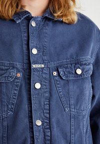 Blå denimjacka med silverfärgade knappar, två bröstfickor och en etikett med texten "REPLAY". Tyget har en mjuk, texturerad yta.
