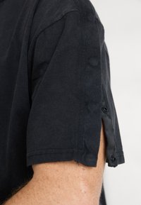 Musto lühikeste varrukatega must särk, millel on külgedel nööpidetail, valmistatud pehmest kangast sileda tekstuuriga. Lihtne, puhas disain.