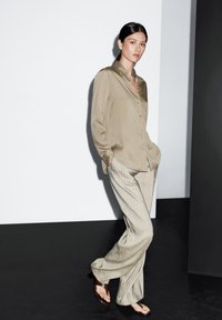 Beige zijden overhemd met knopen, gecombineerd met beige brede geruite broek. Model draagt bruine sandalen; moderne minimalistische achtergrond in grijstinten.