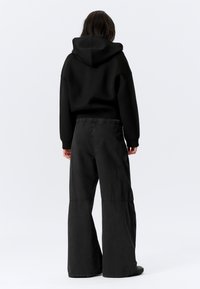 Mustast lõhikuga kapuutsiga dressipluus koos hoogsate, kõrge vöökohaga mustade laia säärega pükstega. Kangas tundub pehme ja mati viimistlusega. Minimaalsed õmbluse detailid.