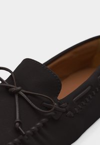 Mocassin en cuir noir avec des détails cousus main et un nœud en lacet noué à l'avant, doublure intérieure couleur tan visible à l'intérieur de la chaussure.