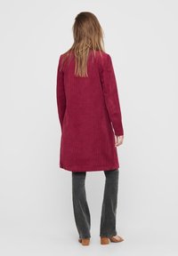 Abrigo largo de pana burdeos con corte recto, bolsillos laterales y abertura trasera, combinado con pantalones grises y calzado marrón.