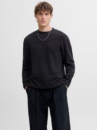 Mladý muž s rukami v vreckách, oblečený v čiernom textúrovanom svetri, striebornej retiazke a čiernych nohaviciach s tenkými pásikmi na jednoduchom pozadí.