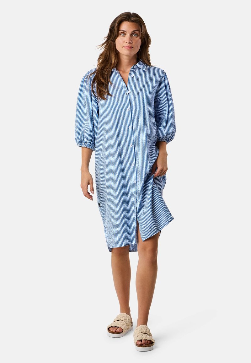 Vestito a righe bleu e bianche con bottoni, dalla vestibilità rilassata, maniche a palloncino e spacco laterale; indossato con sandali beige senza lacci.