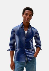 Camisa de botones con rayas azul marino y blancas, fabricada en tejido ligero, con un bolsillo y las mangas remangadas, combinada con unos vaqueros azul claro.