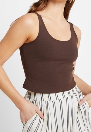 Woman wearing sleeveless dark brown crop top and high-waisted white pants with vertical black stripes, hands in pockets.