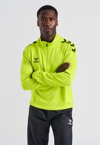 Maglia sportiva gialla fluorescente con zip a metà, logo nero e dettagli neri sulle spalle, abbinata a pantaloni neri. Texture liscia e design aderente.