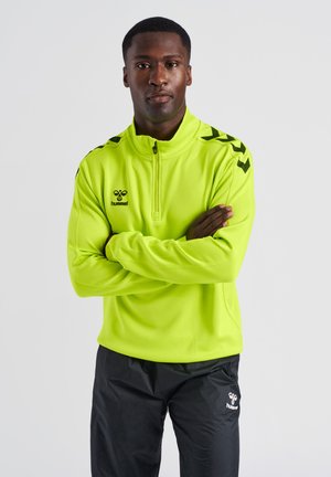 Fluorescerend geel sportief half-zip top met zwart logo en schouderaccenten, gecombineerd met zwarte broek. Soepele textuur en slim fit ontwerp.