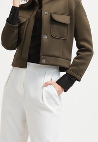 Osoba w oliwkowej, krótkiej kurtce z przednimi kieszeniami, nosząca czarną koszulę, w połączeniu z białymi, plisowanymi spodniami w wysokim stanie, z ręką w kieszeni.