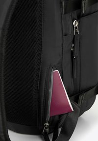 Zaino in nylon nero con pannello a rete, multiple tasche con zip, che presenta un oggetto triangolare viola parzialmente inserito in una tasca.