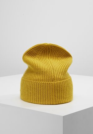 Bonnet en maille côtelée jaune moutarde avec revers plié présenté sur un socle blanc sur un fond gris uni.