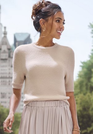 Beiger Pullover mit kurzen Ärmeln, einer strukturierten Oberfläche und rundem Ausschnitt, kombiniert mit einem hellen Faltenrock. Weiche Materialien und dezente Farbtöne.