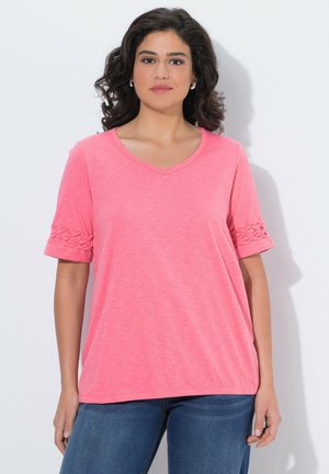 Kobieta o ciemnych kręconych włosach, ubrana w różowy top z krótkim rękawem i dekoltem w serek z plecionymi detalami na rękawach oraz niebieskie dżinsy, stojąca na tle białej ściany.