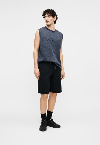 Νέος άνδρας με σγουρά μαλλιά που φοράει αμάνικη σκούρα γκρι μπλούζα Off-White, μαύρο σορτς, μαύρες κάλτσες και μαύρα αθλητικά παπούτσια, στέκεται και κοιτάζει πλάγια.