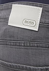 Étiquette de marque rectangulaire blanche avec le texte "BOSS HUGO BOSS" cousue sur la ceinture d’un jean en denim gris foncé.