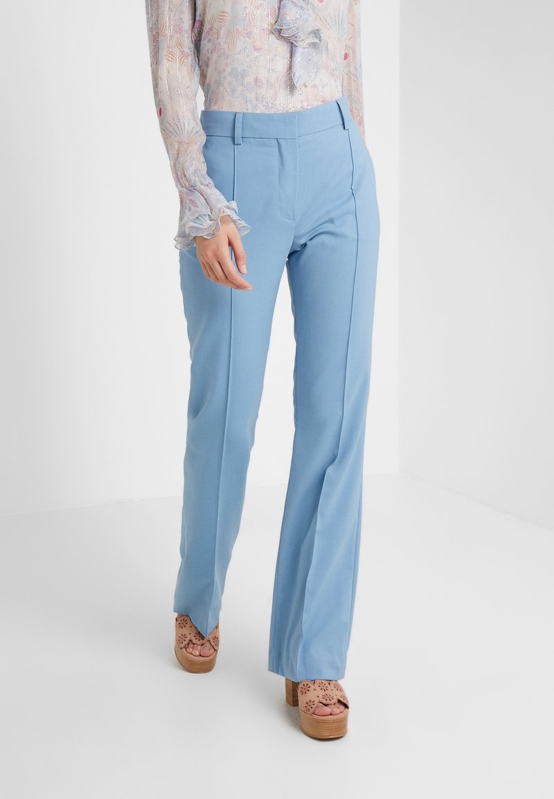 Femme portant un pantalon bleu clair sur mesure et un chemisier transparent à fleurs avec des poignets à volants, associé à des sandales à plateforme beiges.