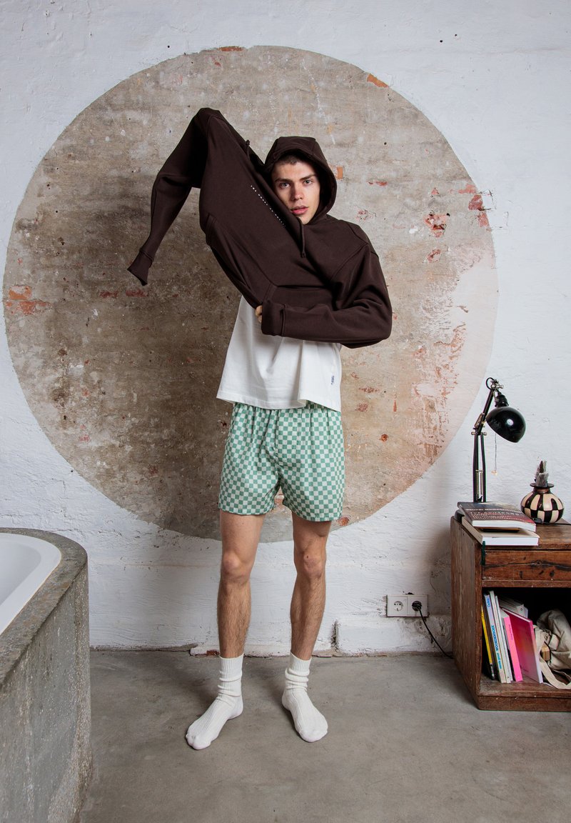 Felpa con zip marrone, camicia bianca e pantaloni corti a scacchi verdi. Il modello si trova davanti a un design murale circolare, indossando calzini bianchi.