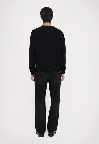 Man standing facing away wearing a black sweater, black jeans, and black shoes against a plain light background.
