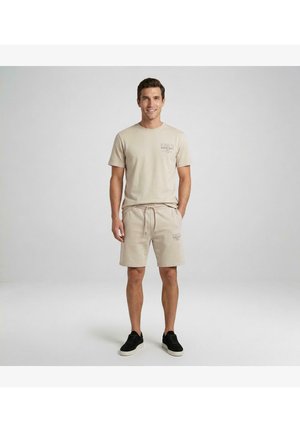 Man staat met een beige t-shirt en bijpassende korte broek met zwarte sneakers tegen een effen lichtgrijze achtergrond.