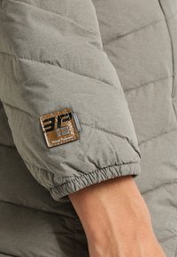 Gray quilted jacket sleeve with elastic cuff, featuring a square brown logo patch with embossed details. Smooth fabric texture.