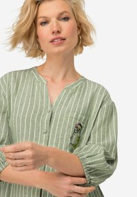 Camicia verde a righe con colletto casual, maniche a palloncino curate e tasca con un design ricamato colorato.