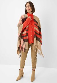 Wollmischung-Poncho mit einem karomuster in Beige, Rosa und Braun, mit fransigen Kanten, kombiniert mit einem roten Sweater und beigen Cordhosen sowie Stiefeln.