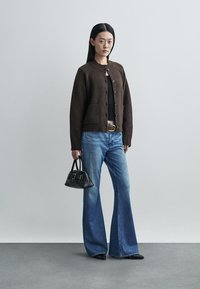 Brown wool cardigan with a round neckline and button closure, paired with blue flared jeans. Black handbag with structured design.