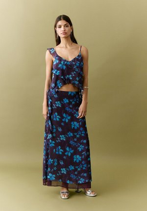 Woman wearing a sleeveless navy dress with blue floral print, ruffled layers, and silver open-toe heels, standing against a plain beige background.