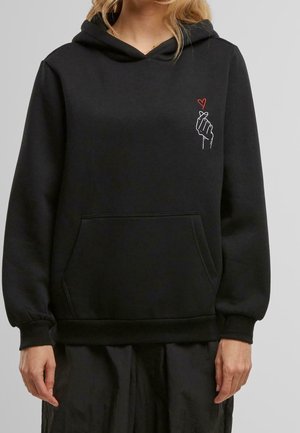 Person iført sort hættetrøje med frontlomme og lille hvid hånd, der laver fingerhjerte med rødt hjerte ovenover på venstre bryst.