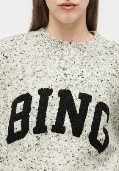 Frau trägt einen grauen, gesprenkelten Strickpullover mit großen, schwarzen, strukturierten Buchstaben, die vorne "BLING" buchstabieren.
