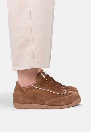 Bruine suède vetersneakers gedragen met lichtbeige broek, met een zijaanzicht van beide schoenen op een witte achtergrond.
