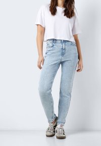 Světle modré džíny s vysokým pasem v kombinaci s bílým zkráceným tričkem. Boty jsou béžové a šedé tenisky. Neutrální pozadí.