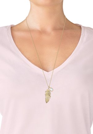 Collana con pendente in oro a forma di foglia, sospesa a una catena sottile, con un ulteriore accento di pietra tonda. Indossata sopra una maglietta rosa chiaro.