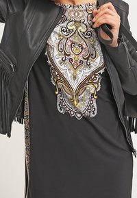 Veste en cuir noir avec des détails à franges et une robe noire à manches longues présentant un motif cachemire coloré et complexe en or, rose et blanc.