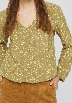 Vrouw draagt een olijfgroene blouse met lange mouwen en witte stippen, en bruine corduroy broek, handen deels in de zakken.