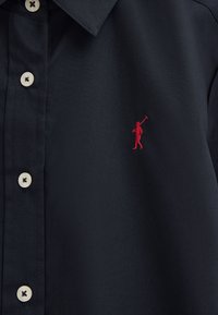 Mørkeblå skjorte med knapper og hvide knapper samt en lille rød broderet logo af en person, der holder en polostav på brystet.