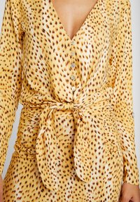 Kvinna i en gul klänning med knappar och bruna leopardfläckar, med ett knytet tygbälte i midjan.