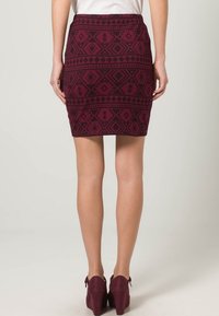 Falda lápiz burdeos con un patrón tribal en negro; diseño ajustado con cinturilla. Combinada con zapatos de tacón oscuros a juego.