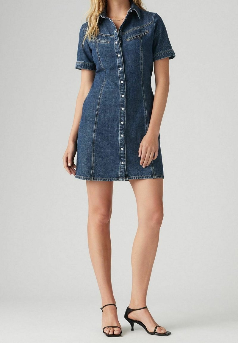 Femme portant une robe en jean à manches courtes, boutonnée sur le devant, avec col et talons ouverts noirs, debout devant un fond gris uni.