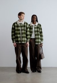 Groene geruite jasjes met witte kunstbontkraag, gedragen over effen tops. Beide modellen dragen bruine wijduitlopende broeken en zwarte schoenen.