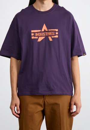 Personne portant un t-shirt ample violet foncé avec une étoile orange et un graphisme texte "Industries" ainsi qu'un pantalon marron, debout devant un fond uni.