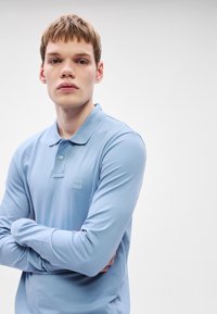 Šviesiai mėlynas ilgomis rankovėmis polo marškinėlis iš minkštos medžiagos, su sagotu apykakle ir mažu kvadratiniu logotipu ant krūtinės.