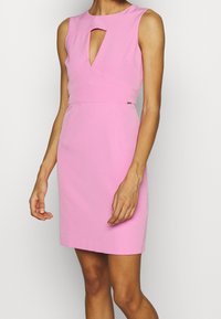 Robe rose sans manches avec un décolleté en forme de goutte, une silhouette ajustée et une texture lisse. Présente des détails de coutures et un petit emblème de marque à la taille.